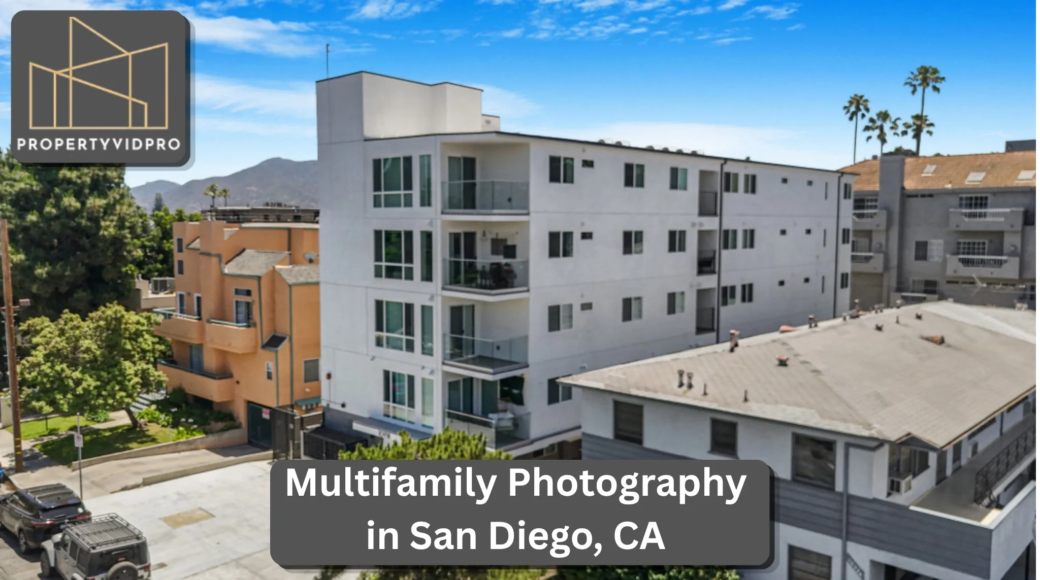 Home 35 Professional aerial real estate photography of a modern, white multi-story apartment building in an urban neighborhood, featuring glass balconies, adjacent properties, parked cars, and distant mountains under a clear blue sky.